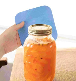 צנצנת זכוכית עם מכסה על משטח עץ, כשברקע יד אוחזת ברפידה כחולה