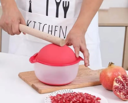 אדם משתמש בפורס רימונים ורוד על קרש חיתוך עץ עם רימונים מסביב